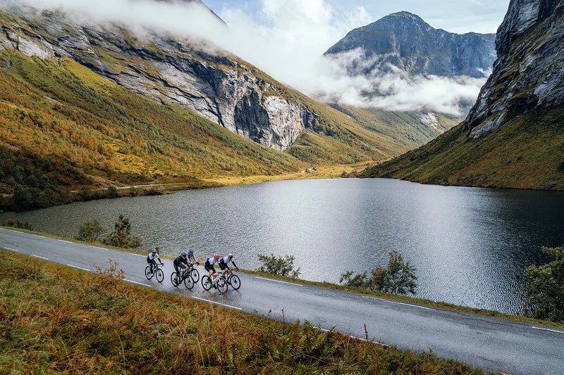 Five cyclists riding along a scenic Norwegian mountain road with a lake and mountains in the background, as part of an organised LeBlanq luxury cycling holiday.