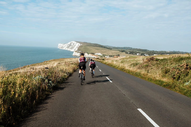 Two cyclists riding on a coastal road with the Isle of Wight’s cliffs in the background.