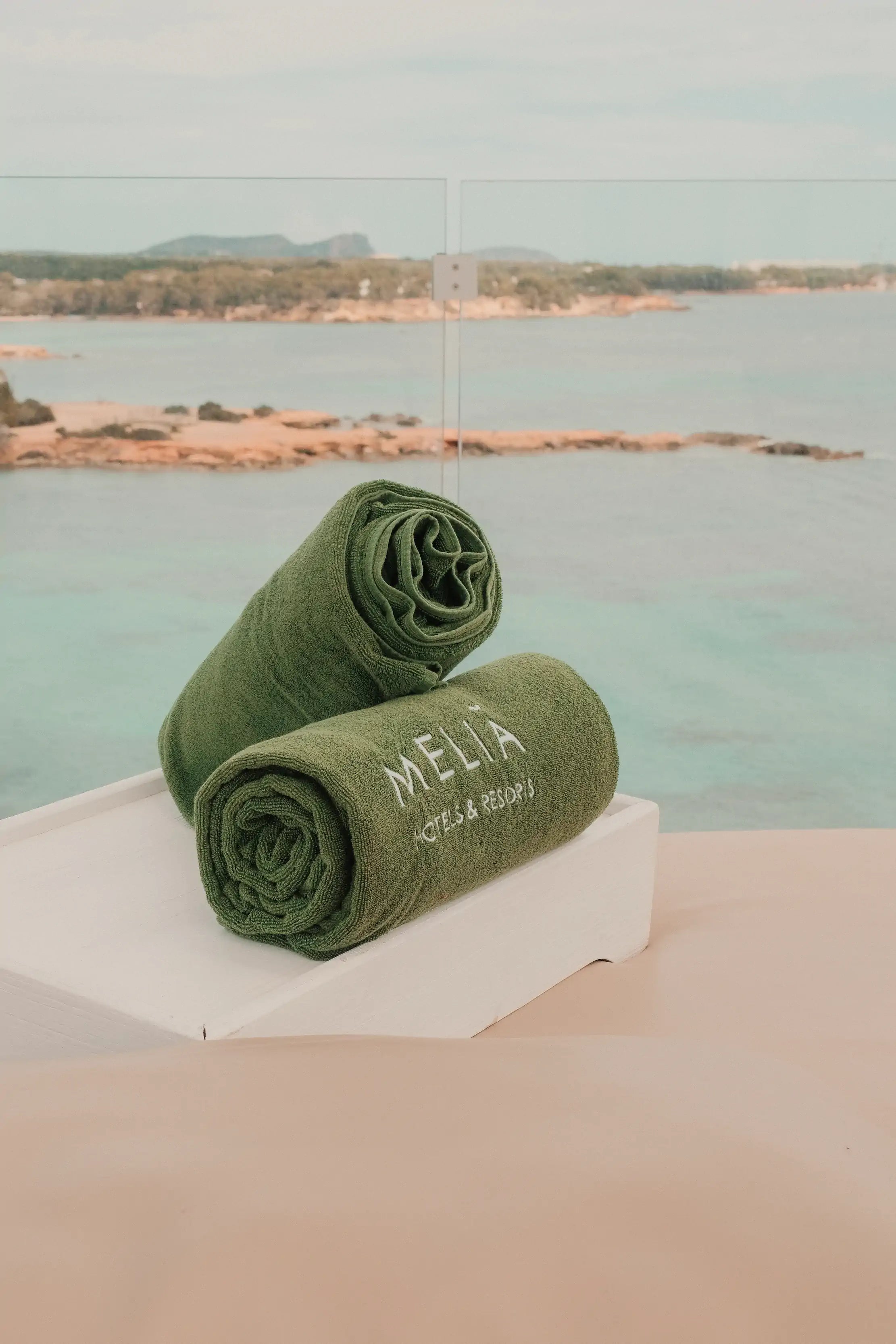 Two green towels with 'Melia' branding on a white block by a body of water.