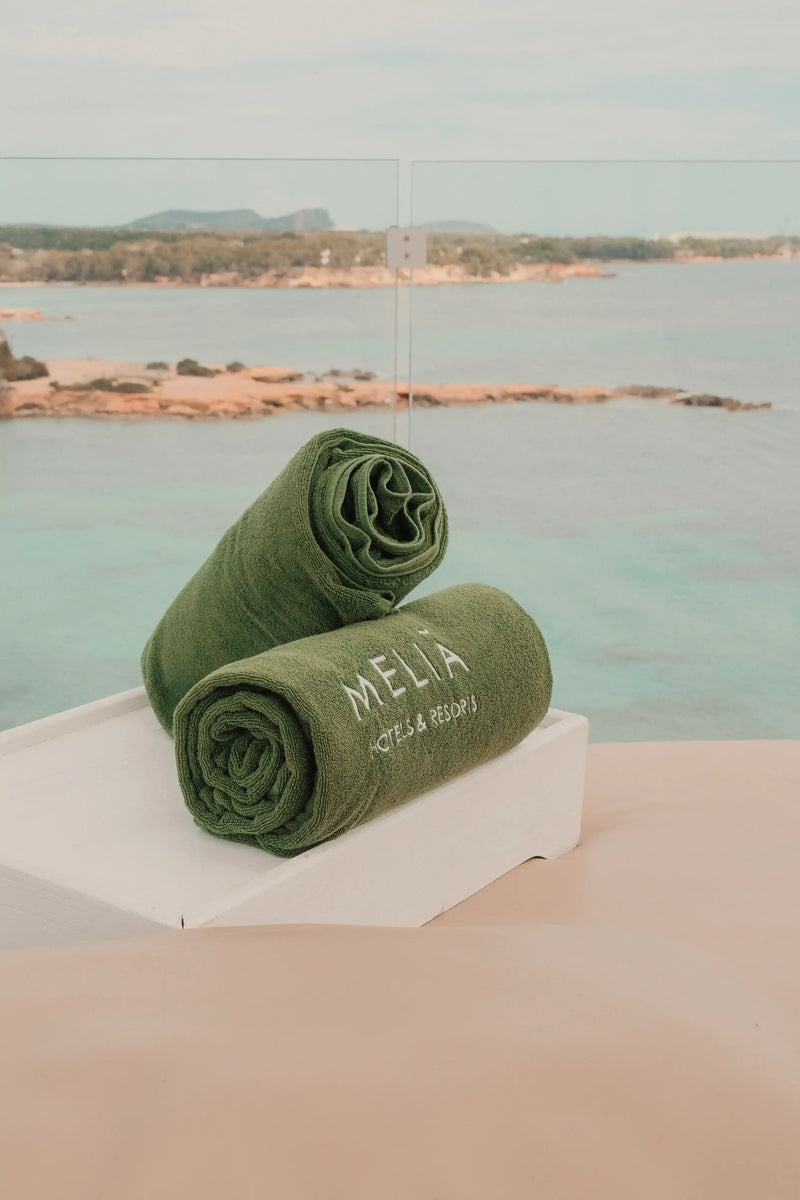 Two green towels with 'Melia' branding on a white block by a body of water.